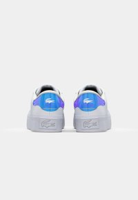 Des sneakers blanches avec une texture lisse, arborant un accent holographique bleu et violet à l'arrière et un logo de marque texturé blanc sur la semelle.