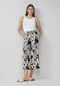 Un débardeur blanc associé à un pantalon large noir et crème à motifs avec des designs de feuilles, comportant des poches et une coupe décontractée. Sandales blanches.