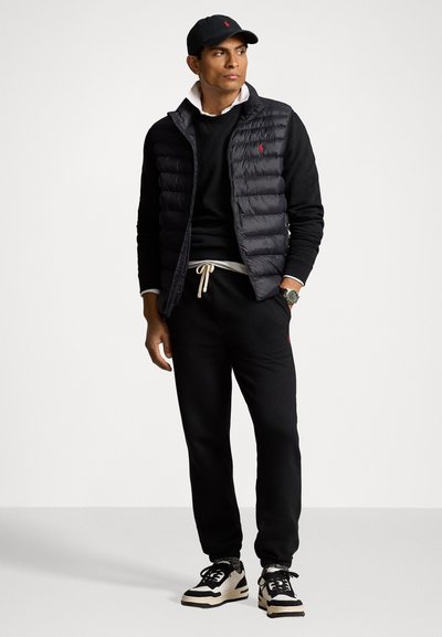 Black puffer vest over a black sweater, white collared shirt, and black joggers. Black cap with logo. Black and cream sneakers.