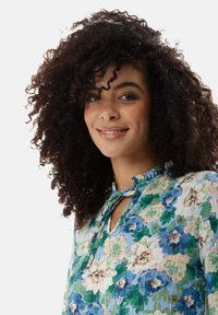 Floral-patterned blouse in light blue with green and pink accents. Features a ruffled collar and tie closure. Lightweight, textured fabric.