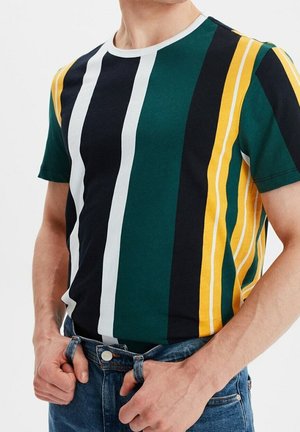 Man draagt een korte mouw T-shirt met verticale strepen in groen, zwart, wit en geel, gecombineerd met blauwe spijkerbroek.