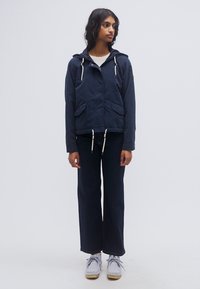 Σκούρο μπλε μπουφάν με κουκούλα, κλείσιμο με κορδόνι και δύο τσέπες μπροστά, σε συνδυασμό με φαρδιά σκούρα παντελόνια και γκρι παπούτσια.