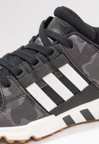 Černá a šedá teniska s maskáčovým vzorem, ozdobená bílými pruhy, texturovanou tkaninou a gumovou podrážkou s černými detaily.