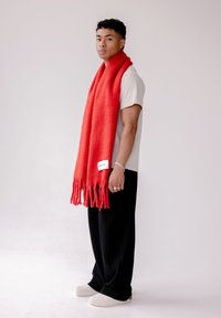 Rode wollen sjaal met franjeranden, over een persoon gedrapeerd die een wit T-shirt en zwarte geribbelde broek draagt, staand op een effen achtergrond.