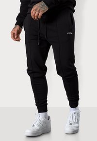Schwarze Jogginghose im Slim Fit, mit elastischem Bund, sichtbaren Nähten, seitlichen Taschen und einem dezenten Logo. Kombiniert mit weißen Sneakern und Socken.