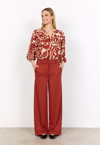 Red wide-leg trousers with a button detail, paired with a cream blouse featuring a bold floral pattern in terracotta and deep red tones.