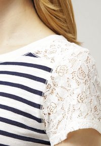 Raidallinen puuvillapaita, jossa on valkoiset pitsiset lyhyet hihat, pyöreä pääntie ja herkät kukkakuviot pitsissä.