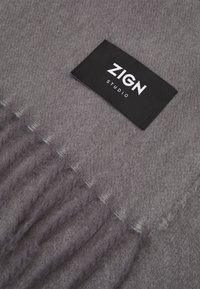 Écharpe grise tricotée avec des bords à franges, comportant une étiquette en tissu noir avec le texte "ZIGN STUDIO" en blanc. Texture douce et matériau chaud.