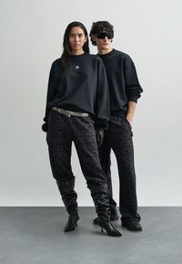 Black oversized sweatshirt with a white emblem, paired with black patterned pants. Model wears knee-high black boots. Detailed textures visible.
