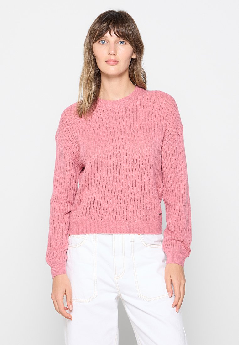 Różowy sweter z dzianiny o prążkowanej strukturze, okrągłym dekolcie i długich rękawach; w połączeniu z białymi spodniami; ma luźny krój.