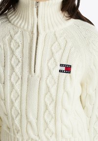 Gros plan d'un pull blanc en tricot torsadé avec un col à demi-fermeture éclair et un patch logo Tommy Jeans sur la poitrine.