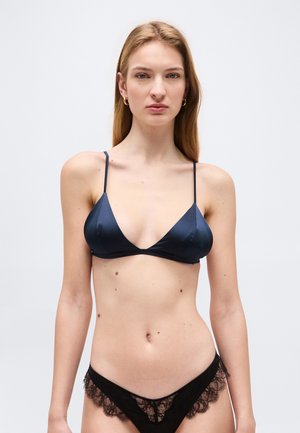 Jonge vrouw die een marineblauwe satijnen bralette en zwart kanten ondergoed draagt, staand tegen een effen lichtgrijze achtergrond.