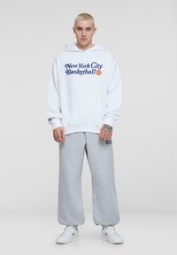 Witte hoodie met de tekst "New York City Basketball" in grafisch ontwerp, gecombineerd met grijze, losse joggingbroek en witte sneakers.