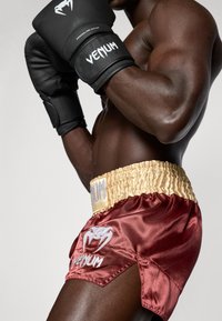 Pantalones cortos de boxeo de satén burdeos con una cinturilla elástica dorada, con un logo blanco y detalles de diseño en el lateral. Guantes de boxeo negros.