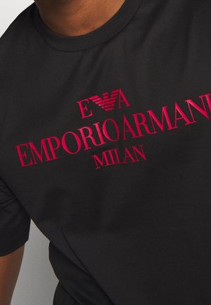 Chemise noire avec logo rouge Emporio Armani Milan et emblème d'aigle sur la partie supérieure de la poitrine, portée par une personne à la peau foncée.