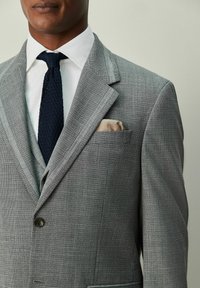 Sacou gri în carouri cu rever ascuțit, închidere cu un singur nasture și o cravată albastru închis contrastantă. Batista din buzunar adaugă un detaliu.