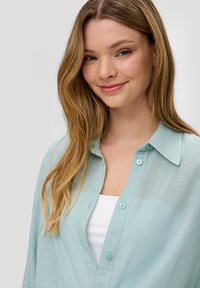 Camicia button-up in lino color turchese chiaro con colletto, realizzata in tessuto traslucido e con bottoni azzurri, indossata sopra una maglietta bianca.