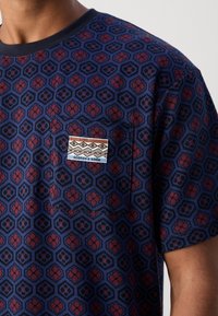 Lyhythihainen, pyöreäkauluksinen paita laivastonsinisessä, jossa on geometrinen kuviollinen kuusikulmioista ja kukista punaisen ja mustan sävyissä; paidassa on rinnassa tasku, jossa etiketti.