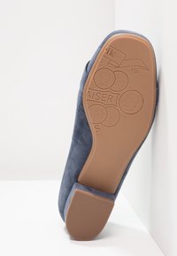 Scarpa piatta in camoscio blu con suola in gomma beige testurizzata. Presenta loghi in rilievo e design di monete sulla suola. Punta arrotondata e tacco basso.