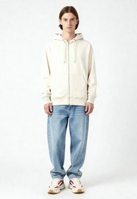 Felpa con zip color crema e cappuccio regolabile, abbinata a jeans denim azzurri chiari. Il modello indossa sneaker bianche con accenti rossi e blu.