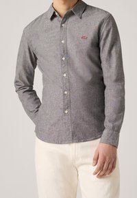 Graues Hemd mit Knopfleiste aus weichem Stoff und langen Ärmeln, mit einem roten Logo-Patch auf der linken Brust, kombiniert mit hellen Hosen.