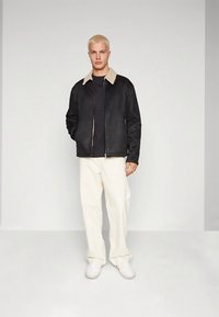 Giacca nera con colletto in shearling beige, chiusura con zip, abbinata a pantaloni bianchi a gamba larga e sneakers bianche su sfondo neutro.