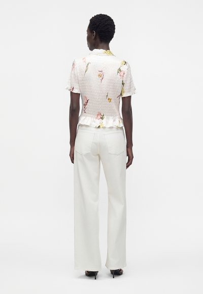 Femme aux cheveux courts portant un chemisier blanc à fleurs à manches courtes et un pantalon blanc taille haute à jambes larges avec des sandales à talons noires.