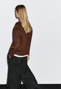 Giacca marrone in misto lana con colletto alto, indossata sopra un top bianco, abbinata a pantaloni a gamba larga a righe nere. Design minimalista, senza chiusure visibili.