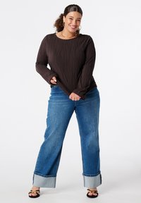 Vrouw die glimlacht in een donkerbruin, gemêleerd langostemshirt, opgestroopte blauwe jeans en zwarte sandalen met hakken en gouden accenten, staande tegen een witte achtergrond.