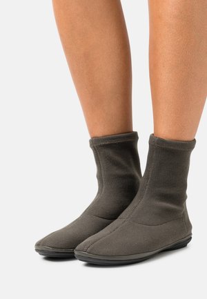 Osoba oblečená v tmavě šedých pletených kotníkových botách se měkkou podrážkou, zobrazená od poloviny lýtek dolů na jednobarevném pozadí.