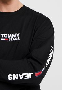 Svart långärmad skjorta i bomull med "TOMMY" och "JEANS" broderat i vitt. Inkluderar färgad logodetalj på ärmen.