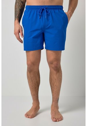 Mann, der leuchtend blaue Badeshorts mit elastischem Bund und Kordelzug trägt, barfuß vor einfachem Hintergrund stehend.