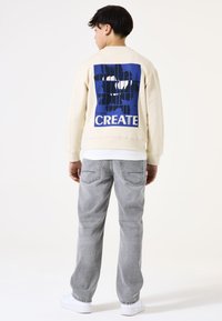 Sudadera crema con un gran gráfico azul que dice "CREATE" en la parte posterior, combinada con jeans gris claro y zapatillas blancas.