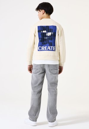 Sweatshirt crème avec un grand graphique bleu portant l'inscription "CREATE" dans le dos, associé à un jean gris clair et des baskets blanches.