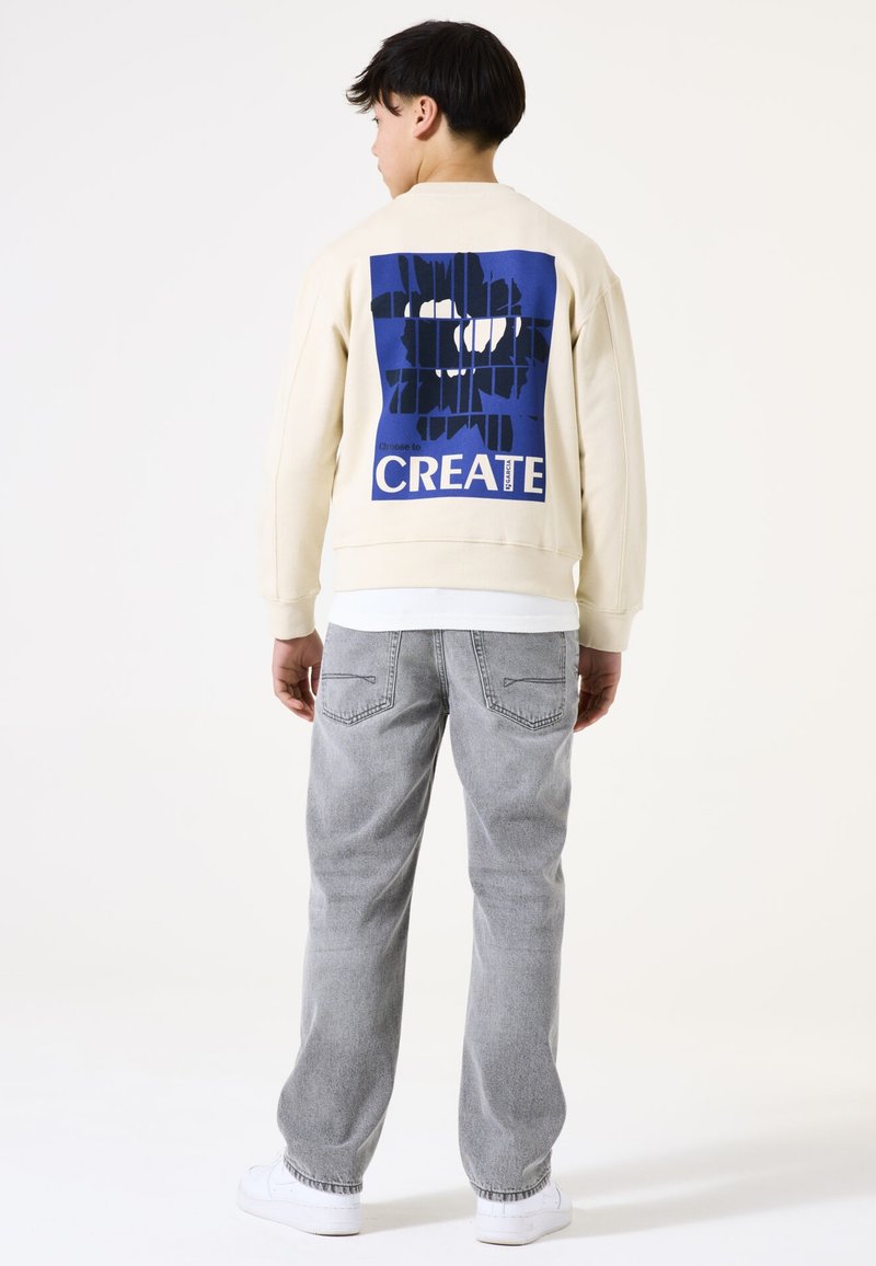 Sudadera crema con un gran gráfico azul que dice "CREATE" en la parte posterior, combinada con jeans gris claro y zapatillas blancas.