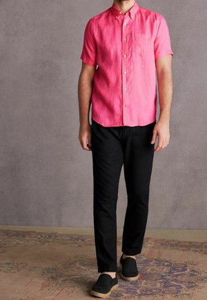 Homme portant une chemise rose vif à manches courtes à boutons, un pantalon noir et des chaussures noires sans lacets, debout sur un tapis à motifs.