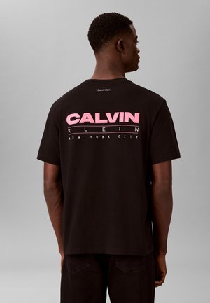 Hombre de espaldas que lleva una camiseta negra de Calvin Klein con la palabra grande “CALVIN” en rosa y un texto más pequeño en blanco que dice “KLEIN NEW YORK CITY” en la parte trasera.