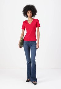 Tricou roșu din bumbac cu decolteu în V asortat cu blugi evazați albaștri și tocuri negre. Modelul ține o poșetă verde, evidențiind o siluetă ajustată.