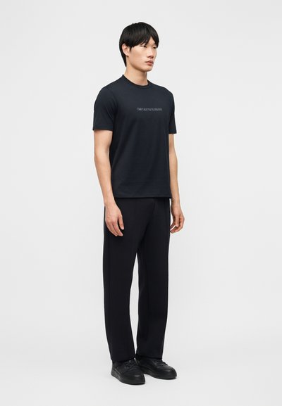 Άνδρας που στέκεται φορώντας μαύρο κοντομάνικο T-shirt με λογότυπο, μαύρο παντελόνι και μαύρα αθλητικά παπούτσια σε απλό ανοιχτογκρι φόντο.