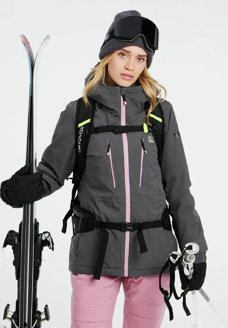 Graue Skijacke mit pinken Akzenten, wasserdichtem Material, verstellbaren Trägern und Reißverschlusstaschen. Kombiniert mit pinken Skihosen und hälftig Skis haltend.
