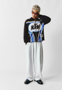 Czarna i niebieska kurtka zapinana na zamek z logo KTM, w parze z szarymi spodniami dresowymi i czarnymi butami z niebieskimi sznurowadłami. Model ma krótkie kręcone włosy.
