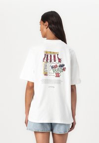 White cotton t-shirt featuring a colorful illustration of a storefront with a striped awning and flower pot on the back. Short sleeves.