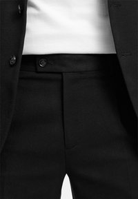Černý oblek na míru s texturovanou tkání, dvěma knoflíky na pase a záhybem na přední straně kalhot. Viditelná bílá košile.