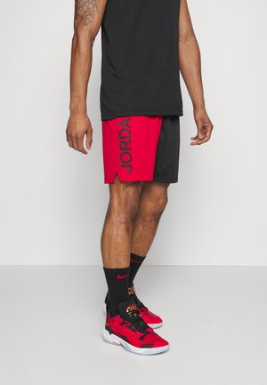 Mężczyzna w czarnej koszulce, czerwono-czarnych spodenkach do koszykówki Jordan, czarnych skarpetkach Nike i czerwono-czarnych sportowych butach stoi w pomieszczeniu.