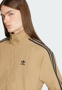 Chaqueta beige con cremallera y cuello alto. Presenta tres franjas negras en los hombros y un logo de Adidas bordado en negro en el pecho. Textura suave.