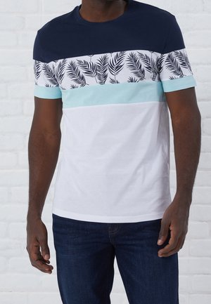 Mężczyzna ubrany w t-shirt z blokami kolorów w poziome paski w kolorach granatowym, białym, jasnoniebieskim oraz z liściastym wzorem, do tego ciemnoniebieskie dżinsy.
