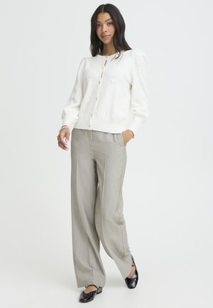 Mujer con suéter blanco abotonado con puntos texturizados, pantalones anchos grises y bailarinas negras, de pie con una mano en el bolsillo.