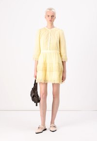 Vestido amarillo con acentos de encaje, dobladillo en capas y mangas tres cuartos. La modelo también lleva un bolso oscuro y usa zapatos de color claro.
