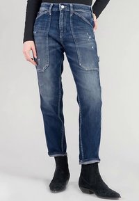 Pantalones cargo de mezclilla azul con un ajuste relajado, que cuentan con bolsillos laterales, detalles desgastados y una pierna ligeramente cónica. Complementados con botas negras.