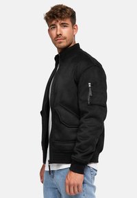 Chaqueta bomber de ante negro con un cuello alto, bolsillos con cremallera, puños acanalados y una textura suave. El modelo lleva una camiseta blanca debajo.